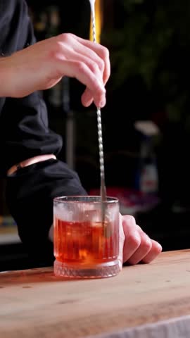 A bartender's hands stir an alcoholic drink with ice using a spoon in a close-up shot. Vertical slow-motion video format, perfect for cocktail preparation and beverage content.
