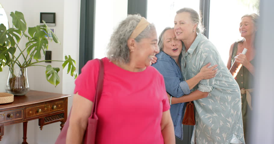 Senior friends share laugh indoors. A biracial woman in pink, a Caucasian in blue, Asian in gray, and a Caucasian in a patterned shirt, all with gray hair, unaltered, slow motion
