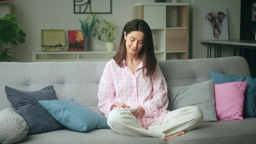 Woman peacefully writes in a notebook on a cozy couch in a well-decorated living room with colorful pillows and plants. 4K video, slow motion, 50 fps