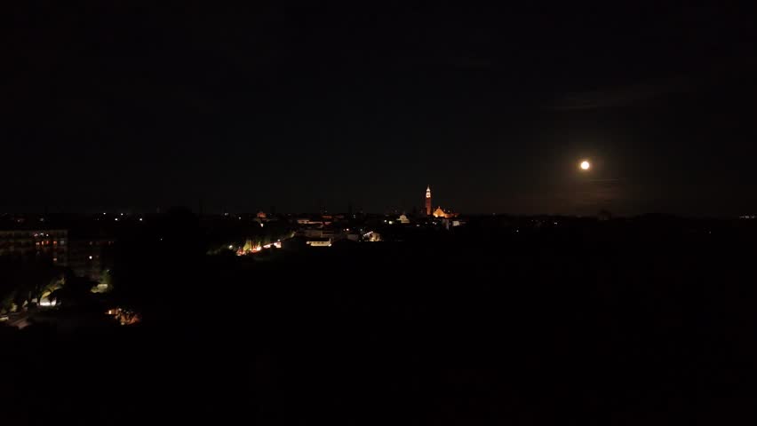 Aerial panoramic view of Cremona city park at night showing illuminated walkways, torrazzo tower and the moon rising in the distance