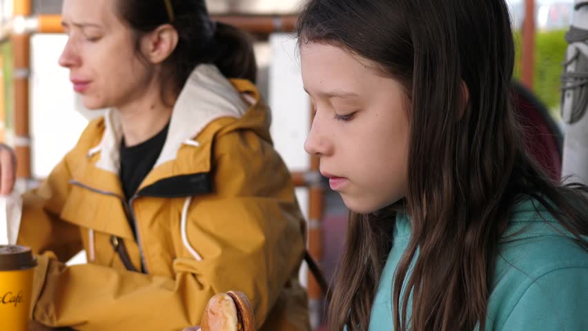 Mother with kid daughter enjoy eating chewing a cheeseburger at outdoor fast food restaurant patio table