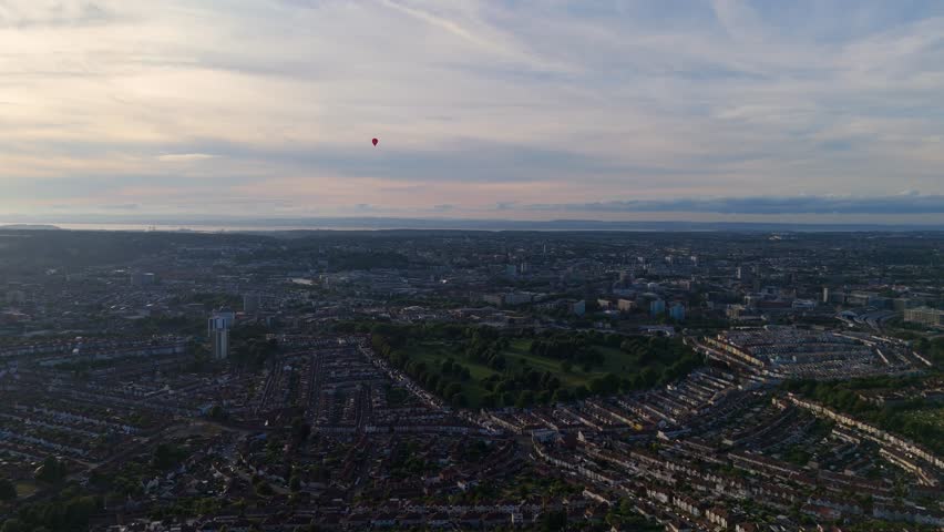 Bristol Ballon Fiesta in late afternoon, golden hour, summer. Dozens of hot air balloons flying over the cityscape horizon. Filmed with 4K drone
