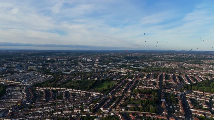 Bristol Ballon Fiesta in late afternoon, golden hour, summer. Dozens of hot air balloons flying over the cityscape horizon. Filmed with 4K drone