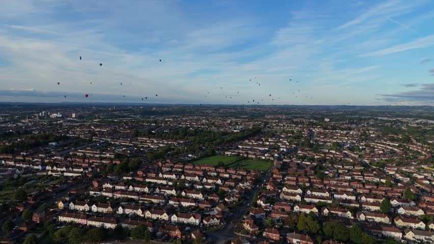 Bristol Ballon Fiesta in late afternoon, golden hour, summer. Dozens of hot air balloons flying over the cityscape horizon. Filmed with 4K drone