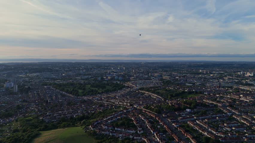 Bristol Ballon Fiesta in late afternoon, golden hour, summer. Dozens of hot air balloons flying over the cityscape horizon. Filmed with 4K drone