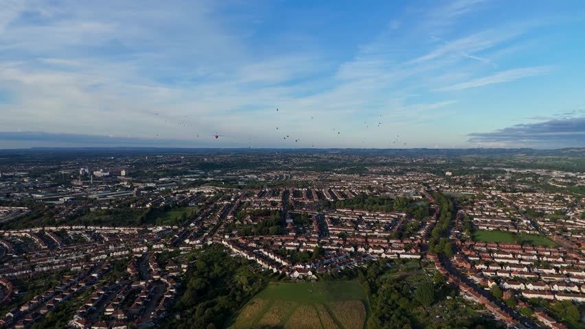 Bristol Ballon Fiesta in late afternoon, golden hour, summer. Dozens of hot air balloons flying over the cityscape horizon. Filmed with 4K drone