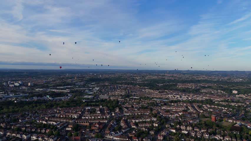 Bristol Ballon Fiesta in late afternoon, golden hour, summer. Dozens of hot air balloons flying over the cityscape horizon. Filmed with 4K drone