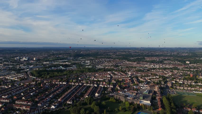 Bristol Ballon Fiesta in late afternoon, golden hour, summer. Dozens of hot air balloons flying over the cityscape horizon. Filmed with 4K drone
