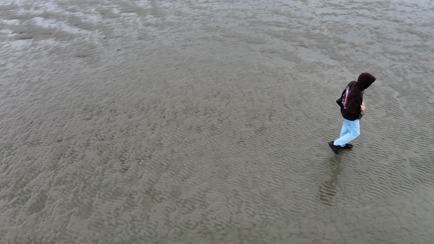 Drone footage of a young man standing on the North Sea seabed during low tide in Cuxhaven, Germany. The video captures the vast sandy expanse.