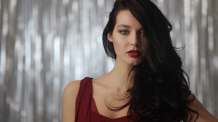 A glamorous woman with long dark hair poses in a red dress against a silver backdrop, exuding elegance and confidence. Portrait of a woman with long dark hair, posing against a shimmering silver