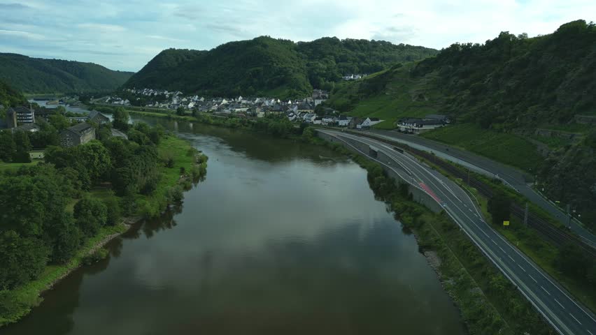 A drone flying over Moselle River towards Kraftwerk Lehmen, surrounded by houses and green mountains in Niederfell, Germany