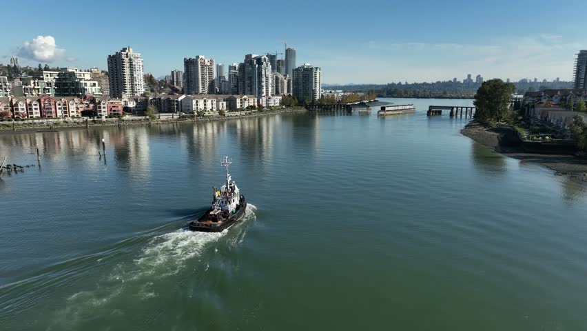A drone view of a Tugboat sailing along Fraser River, on a sunny day with New Westminster buildings in the background in British Columbia, Canada