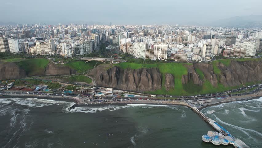 A drone flying above the sea, showing a road near the green coast Peruvian beach in the bay of Lima, with city on cliff in the background in Peru