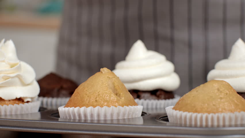 A person pipes frosting onto freshly baked cupcakes in a home kitchen.