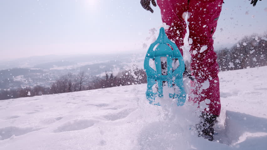 SLOW MOTION, LENS FLARE, LOW ANGLE, CLOSE UP: Icy snowflakes fly in the air as playful female wearing snowshoes runs in deep snow. Energetic young woman runs across the wintry landscape in snowshoes.