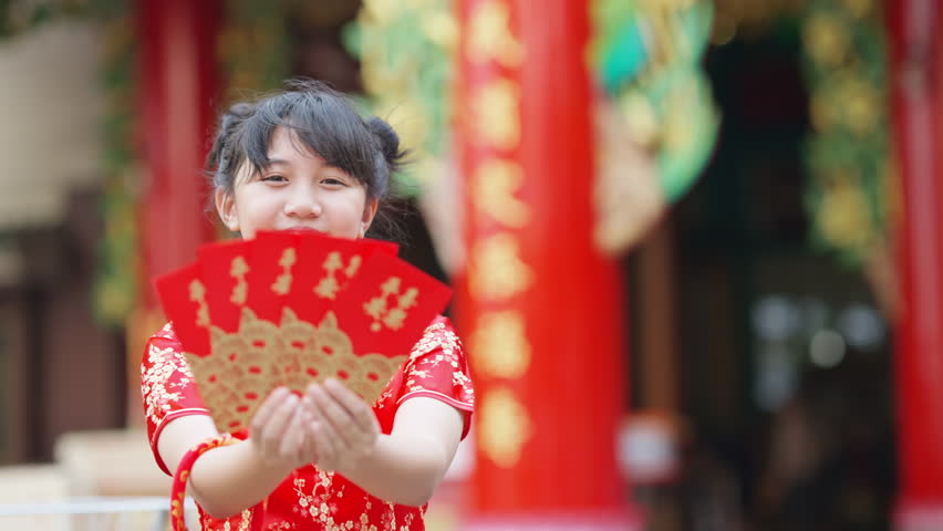 Happy Little Asian girl in traditional Chinese red dress holding red envelopes with money for good luck standing in front of Chinese shrine. Chinese lunar new year holiday festival celebration concept