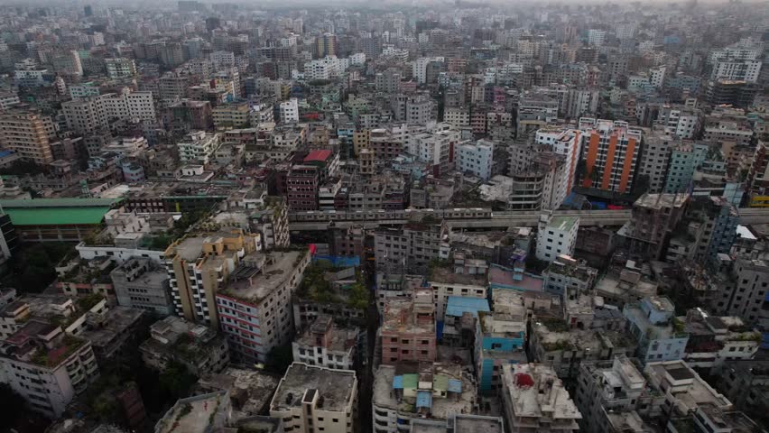 An aerial footage of Dhaka Metro Rail Train (DMRT) moving on a flyover rail track through a forest of urban buildings at sunset in Dhaka, Bangladesh