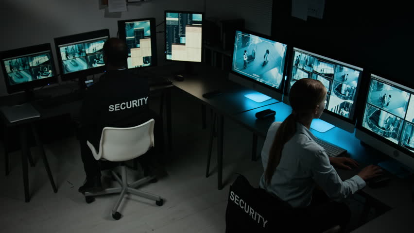 Full high-angle shot of two multiethnic security officers working in control room, Caucasian female guard noticing suspicious activity on CCTV cameras, discussing with African American male colleague
