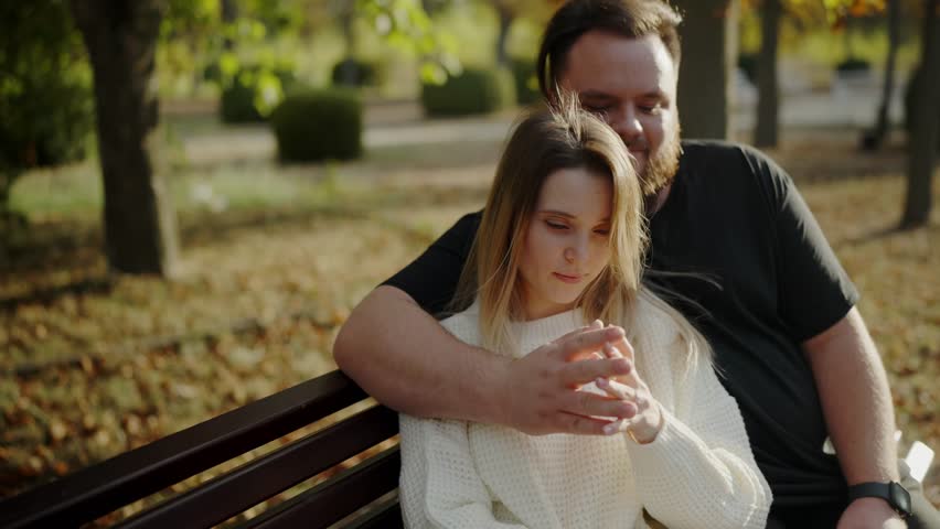 A young couple sitting on a park bench, entwined hands and enjoying a peaceful, intimate moment. The setting sun adds a warm glow, emphasizing affection and togetherness. Intimate Couple