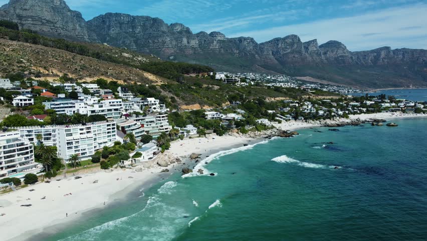 A drone flying over Clifton Beach showcasing a blue seascape, white sand and mountainous buildings in Cape Town, South Africa