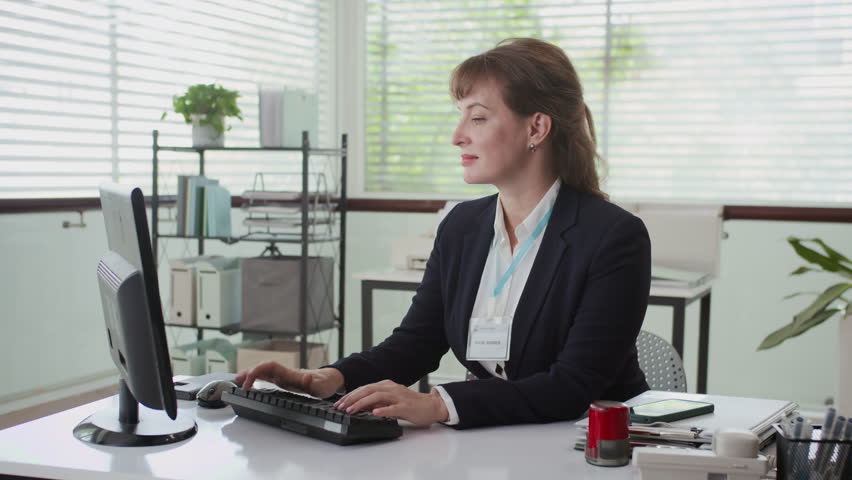 Medium portrait of female Caucasian manager in formalwear smiling at camera while working at office desk