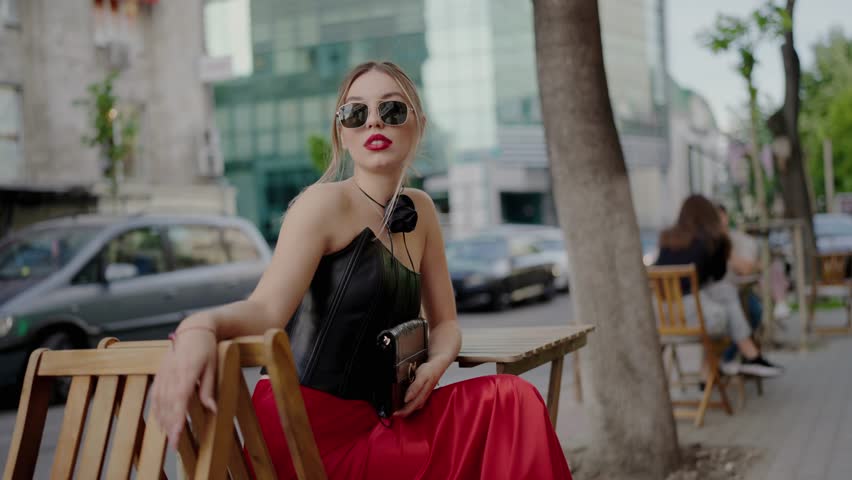 Young fashionable woman wearing sunglasses and leather top sitting at an outdoor cafe in the city, looking through her purse. Urban lifestyle and street fashion. Fashionable Young Woman in Sunglasses