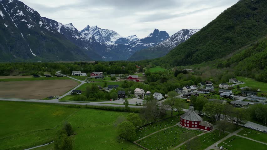 An aerial view of the red Rauma Church and cemetery among green trees, surrounded by snowy mountain tops in Rauma, Norway