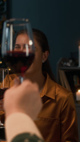 Vertical slowmo shot of several cheerful friends raising glasses of red wine and toasting during cozy Christmas dinner party at festive table in cozy warm lit room decorated with tree and candles