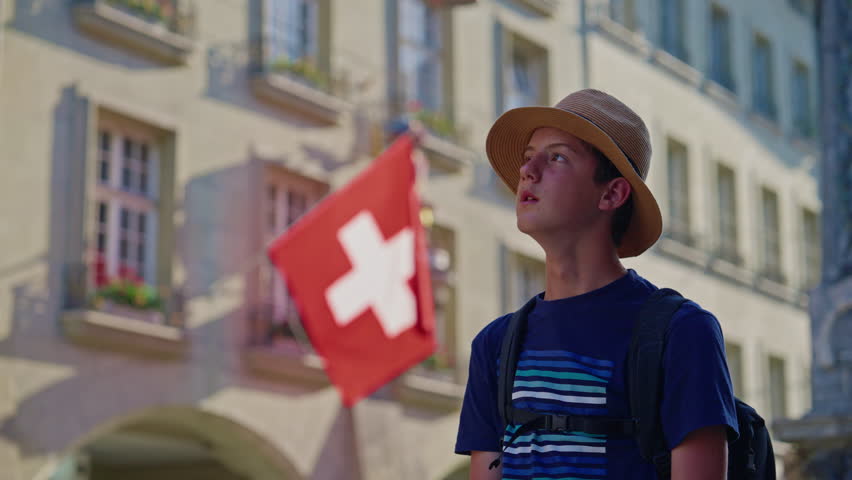 Young guy student tourist with backpack and hat in the center of the old city of Bern watching architecture, buildings around on the background of the Swiss flag. Switzerland