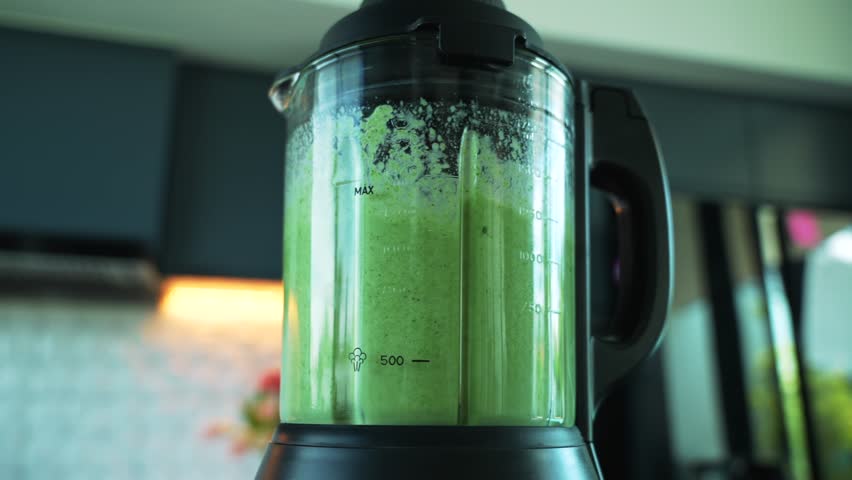 Close-up of transparent glass electric blender bowl making green smoothie in kitchen