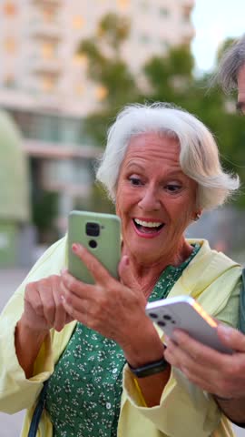 Vertical close-up photo of a caucasian happy modern senior couple using smartphone in the city