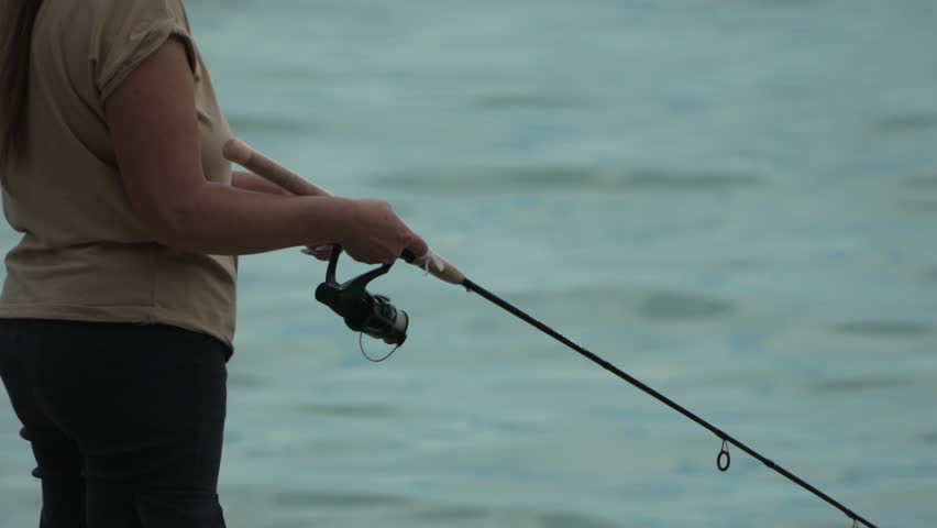 Woman fishing hobby on sea tightens a fishing line reel of fish summer. Calm surface sea. Close-up of a fisher lady hands twist reel with fishing line on a rod. Rock fishing in sea outdoors. Slow