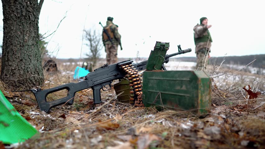 On the ground in a field there is a light machine gun with cartridges against the background of soldiers. Close-up of machine gun cartridges. Small automatic weapons. Light machine gun. Firearms