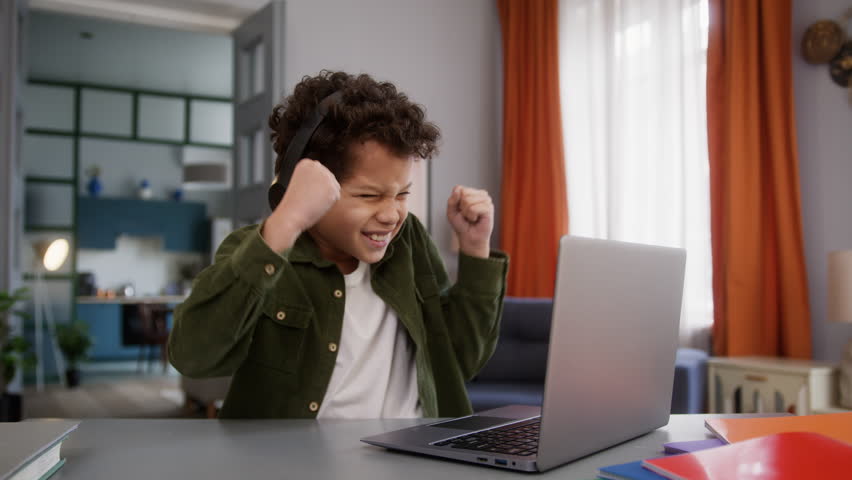 Funny little African American boy son kid child gamer at home table playing online video game internet gaming laptop computer win success winner victory wearing headphones achieve celebrate triumph - Powered by Shutterstock - Get 15% off with code: PIKWIZARD15