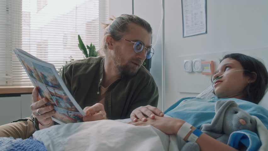 Side slowmo shot of Caucasian dad in glasses reading comic strip to little sick daughter with nasal cannula and bunny plush lying in hospital bed during recovery period in clinic