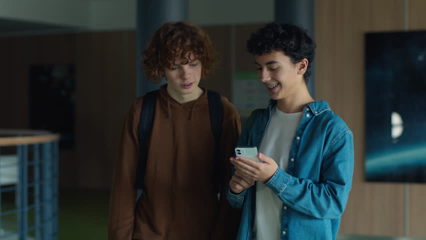 Two teenagers friends Caucasian males men guys pupils students schoolboys walking going in corridor university college school boys looking in mobile phone app shocked laugh smile talking online news