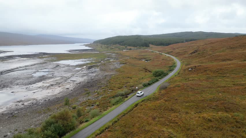 distant white suv driving along remote road scottish highlands beautiful lake river background picturesque drone shot tracking car scotland hills aerial adventure nature landscape travel