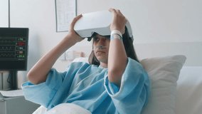Side shot of little girl with nasal cannula putting on VR headset and observing hospital ward in metaverse, lying in bed during recovery period in child health centre - Powered by Shutterstock - Get 15% off with code: PIKWIZARD15