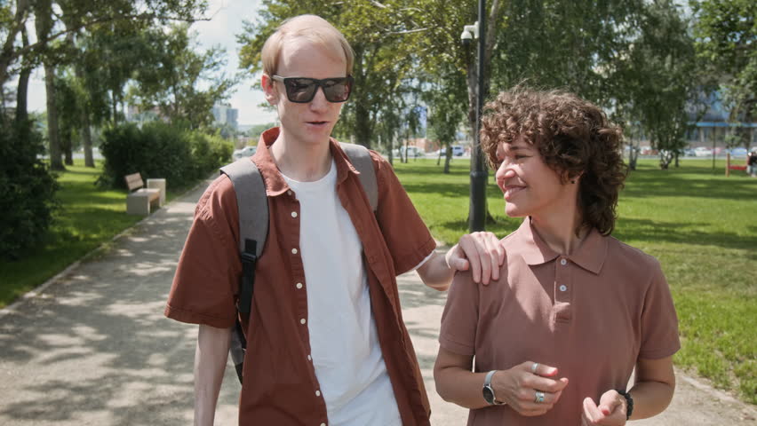 Tilt down view of two diverse friends walking and chatting in park on summer day, visually challenged man with cane holding hand on womans shoulder