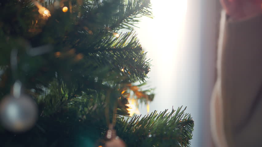 Close-up hand of woman decorating Christmas tree with baubles balls on the background of festive lights at home on calm winter evening, Christmas and new year concept.