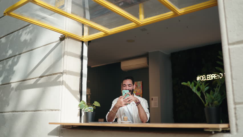 Hispanic adult man sitting by open café window, using his smartphone while sipping coffee. Sunlight pours in, creating relaxed atmosphere. Focus on technology, connectivity, and remote communication - Powered by Shutterstock - Get 15% off with code: PIKWIZARD15