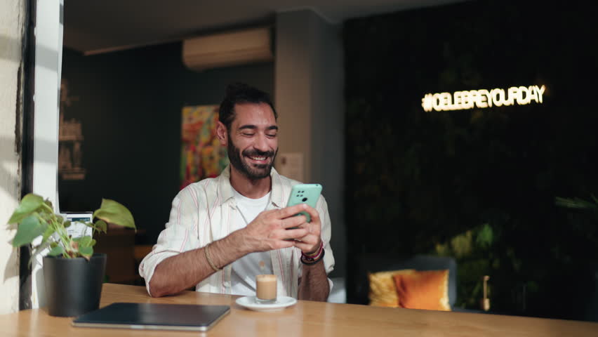 Smiling man scrolling through social media on smartphone device enjoying coffee in cozy café. Casual and relaxed setting with focus on technology and online engagement. Fast 5G internet connection - Powered by Shutterstock - Get 15% off with code: PIKWIZARD15