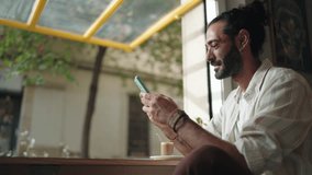 Hispanic man sits by café window, smiling as he checks his smartphone app, engaging in online banking or mobile transactions. Sunlight filters through, creating relaxed and cozy atmosphere - Powered by Shutterstock - Get 15% off with code: PIKWIZARD15