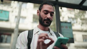 Hispanic man waiting at bus stop, checking public transport schedule on smartphone. Male tourist focused and prepared, using mobile app to stay informed about arrival times in urban setting - Powered by Shutterstock - Get 15% off with code: PIKWIZARD15
