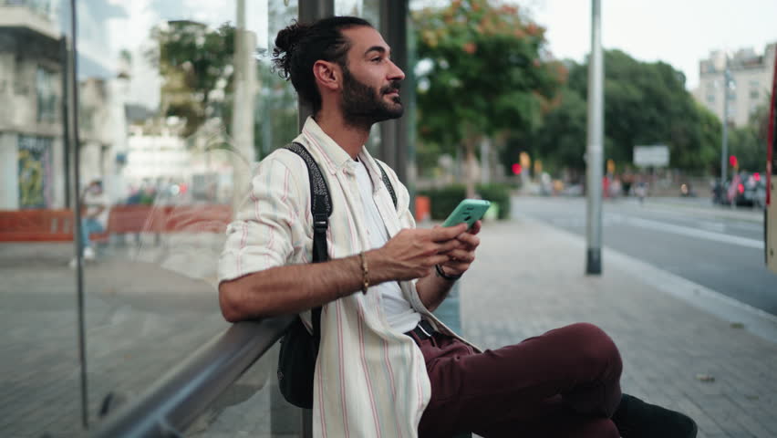 Male tourist using smartphone at bus stop, checking navigation or transport schedule. Dressed casually with backpack, man appears focused, utilizing digital tools for convenient travel planning - Powered by Shutterstock - Get 15% off with code: PIKWIZARD15