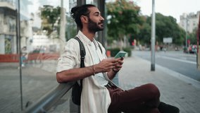 Male tourist using smartphone at bus stop, checking navigation or transport schedule. Dressed casually with backpack, man appears focused, utilizing digital tools for convenient travel planning - Powered by Shutterstock - Get 15% off with code: PIKWIZARD15