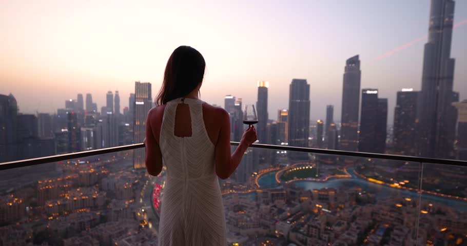 A beautiful tourist woman in a white dress holding a drink enjoys the evening view behind the modern skyline of Downtown Dubai, UAE