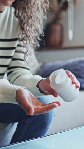 Woman sitting on a couch opens a pill bottle and prepares to take her medication for health and wellness.   Woman taking medication from pill bottle