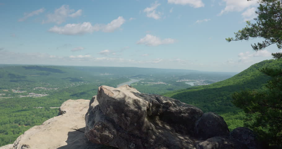 Young man successfully reaches top of a mountain after long climb. Stands confidently and proudly on a rock overlooking the amazing view and natural landscape. Green plains and forest trees and river