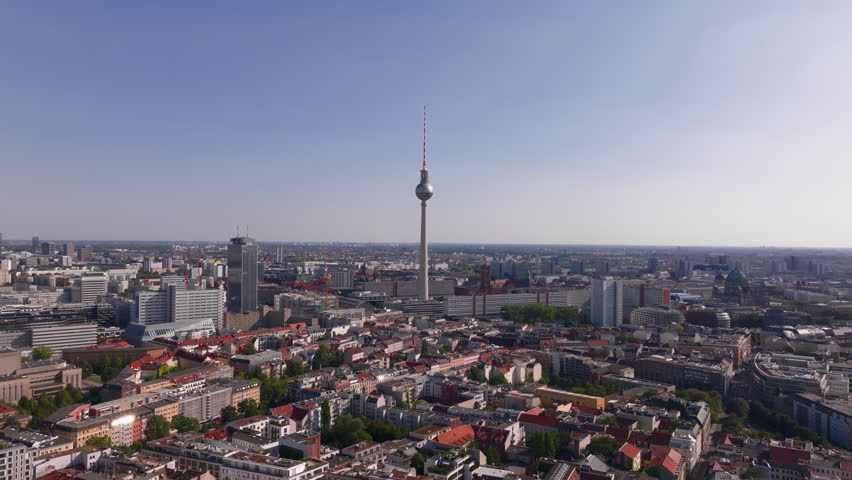 Aerial views showcase the iconic Berlin tv tower in Alexanderplatz rising above a vast cityscape, illuminated by warm sunlight. Expansive buildings and streets stretch endlessly in every direction